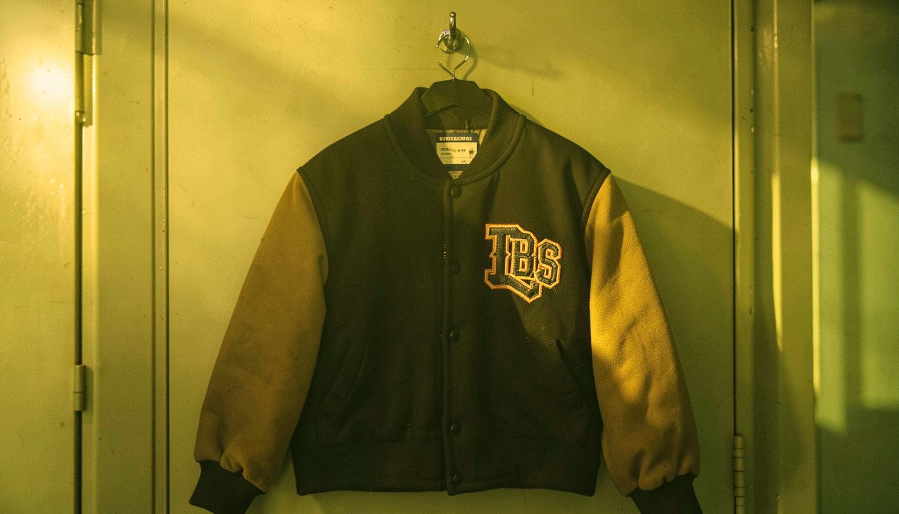 A classic college jacket with contrasting sleeves and embroidered lettering, hanging on a locker room hook in golden light.