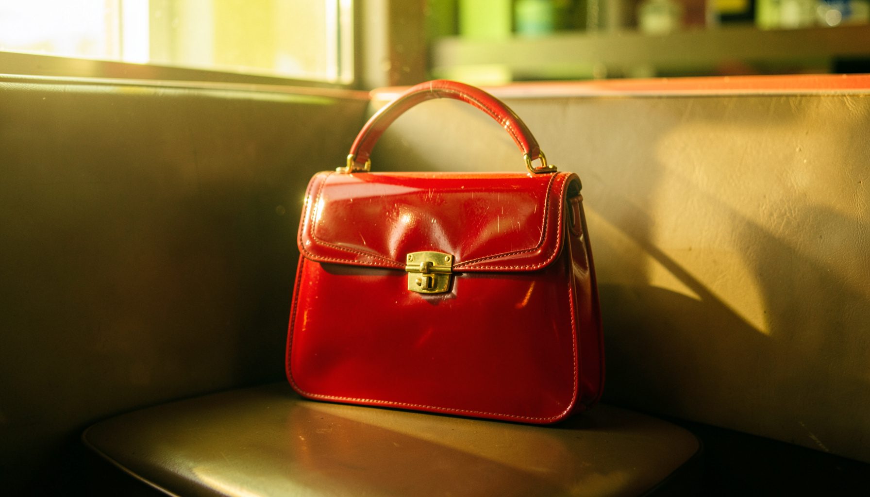 A vintage-style handbag in red patent leather with a gold clasp, resting on a diner booth seat in afternoon light.